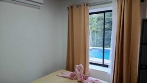 Quarto com cama, cortinas bege e janela com vista para uma piscina.