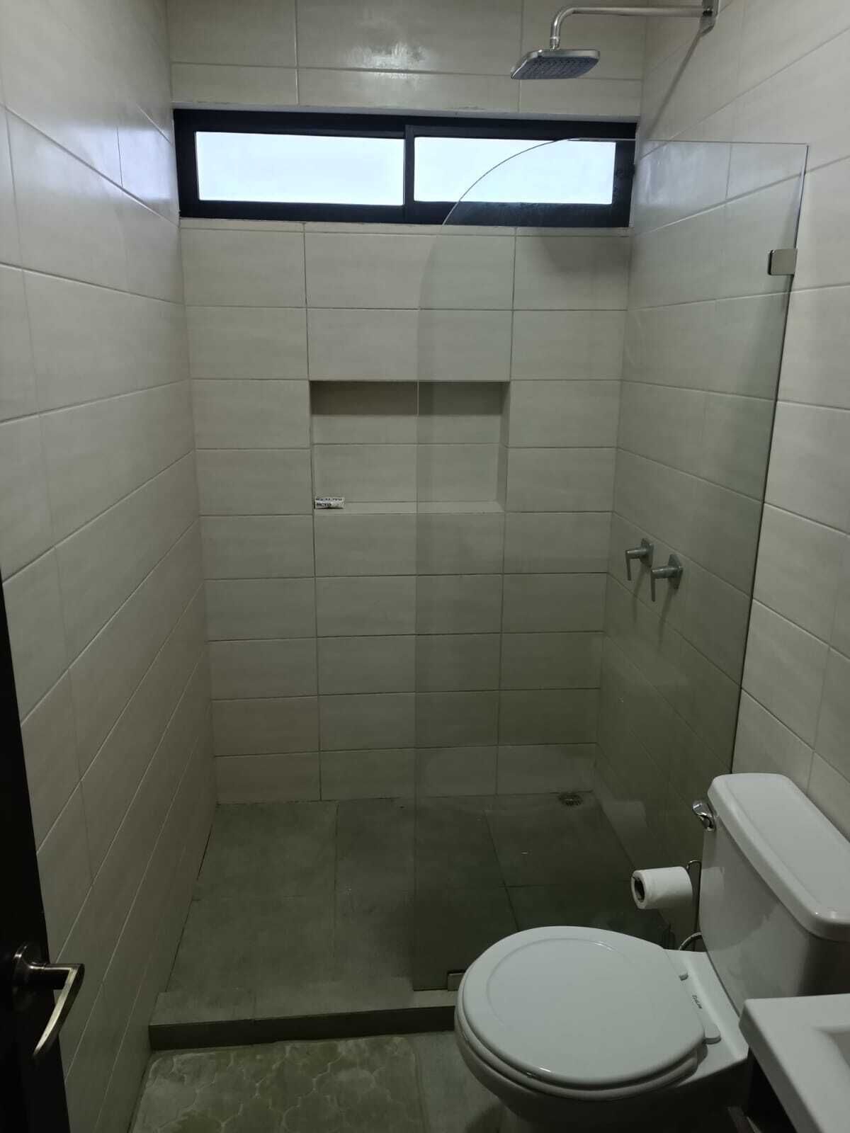 A small, tiled bathroom with a shower, a toilet, and a window above the shower.