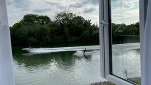 Une vue depuis une fenêtre donnant sur une étendue d’eau, avec un pratiquant de wakeboard, des arbres et une zone herbeuse où se trouve une borne d’incendie.
