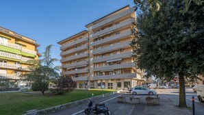 Un edificio residencial de varios pisos con balcones, un espacio pavimentado con una moto estacionada y una calle bordeada de árboles.