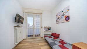 Una habitación con cama, radiador, televisor, ventana con cortinas y un cuadro colorido en la pared.
