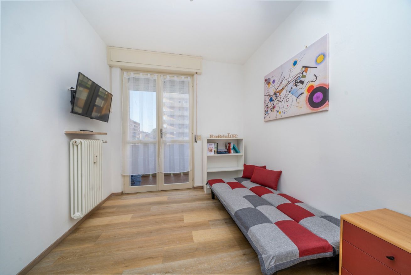 A room with a bed, a radiator, a TV, a window with curtains, and a wall with a colorful painting.
