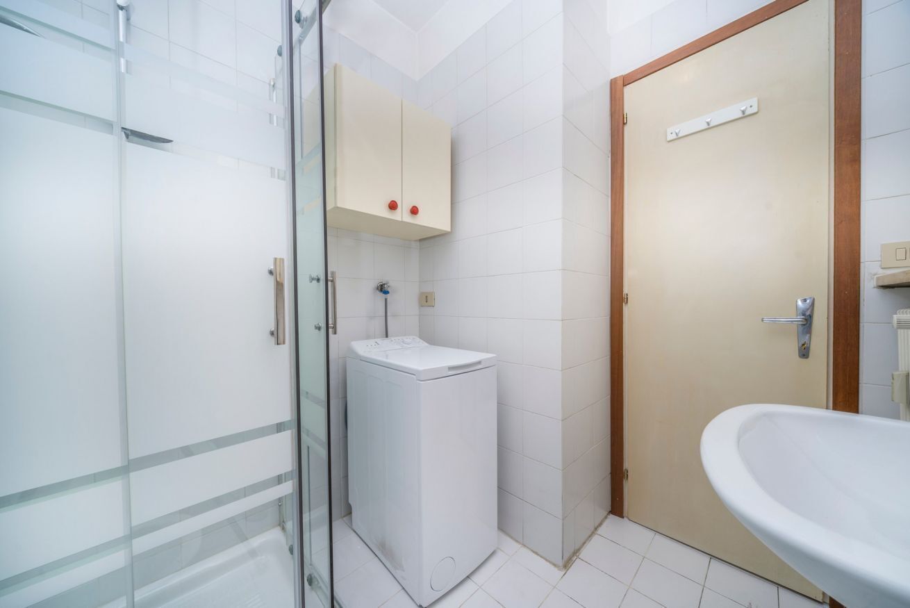 A bathroom with a glass shower enclosure, a washing machine, a wooden door, and a sink.
