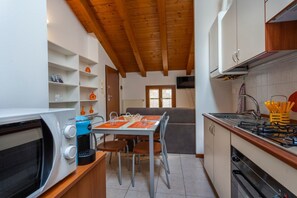 Cocina compacta con microondas, cafetera y estufa. Hay una mesa de comedor con sillas y un banco. La cocina tiene vigas de madera en el techo y armarios blancos.