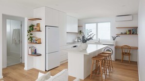 A modern kitchen with white cabinetry, a central island with stools, wooden shelves with books and plants, and a window providing natural light. - The Breeze - holiday apartment in Merewether, Newcastle (Merewether)