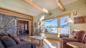 Un interior acogedor con piso de madera, un sofá, una silla y una mesa. Un ventanal ofrece vistas a un paisaje nevado.