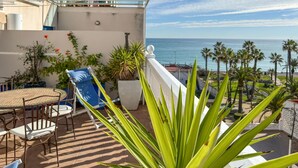 Un balcon avec une table et des chaises, offrant une vue sur la côte, avec des palmiers et un ciel dégagé.