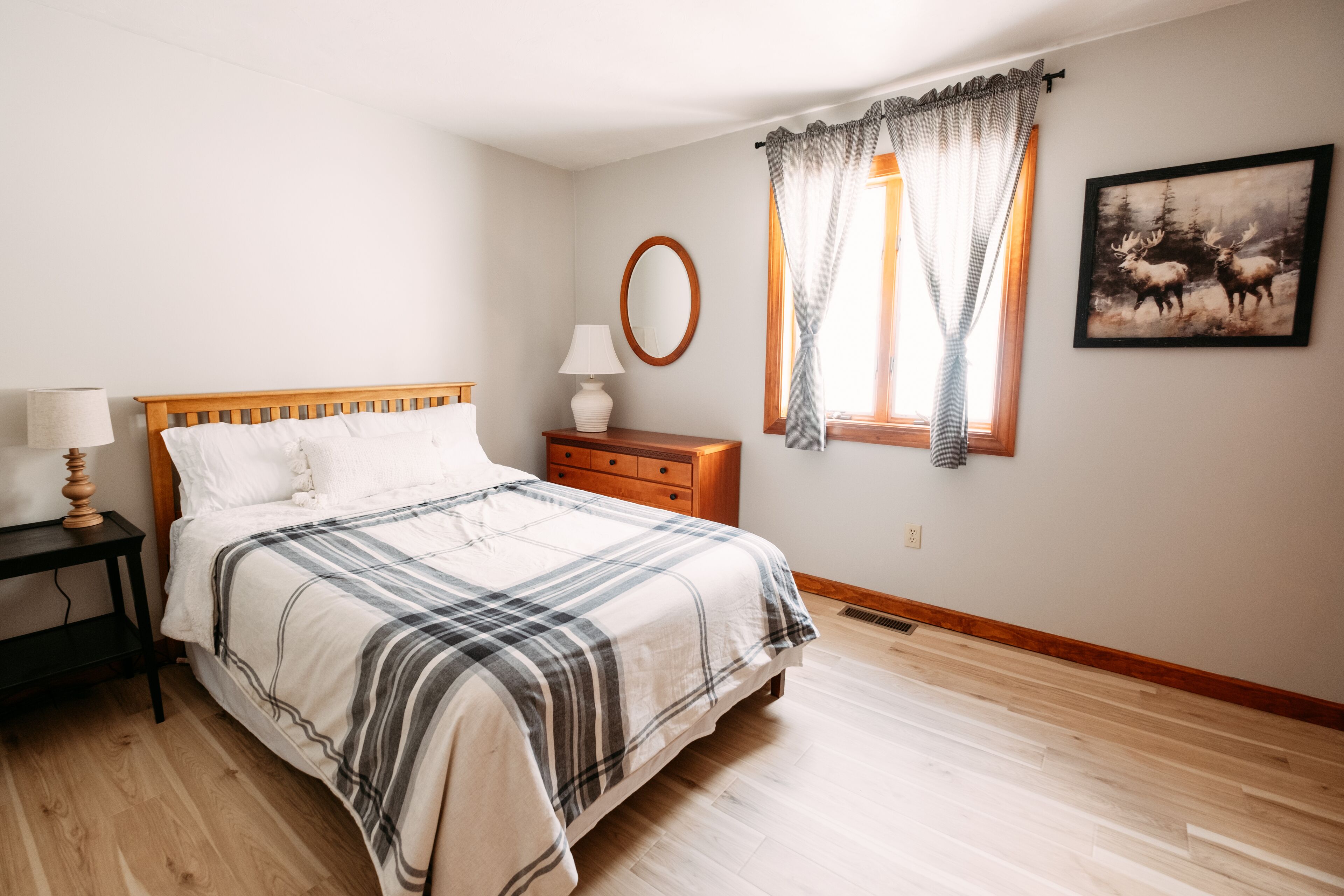 Un dormitorio con cama, mesita de noche, lámpara, cómoda, ventana con cortinas y un espejo adosado a la pared.
