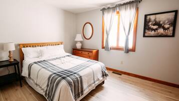 Un dormitorio con cama, mesita de noche, lámpara, cómoda, ventana con cortinas y un espejo adosado a la pared.