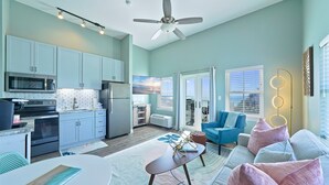 Un salon moderne avec un canapé bleu, une table basse blanche et un coin cuisine doté de meubles bleu clair et d’appareils électroménagers en acier inoxydable.