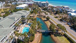An aerial view of a coastal hotel with a swimming pool, palm trees, and a bridge over a water channel. - WOW! Water View Corner Unit, Free Wine + Bottled Water Arrival Gift (Santa Rosa Beach)