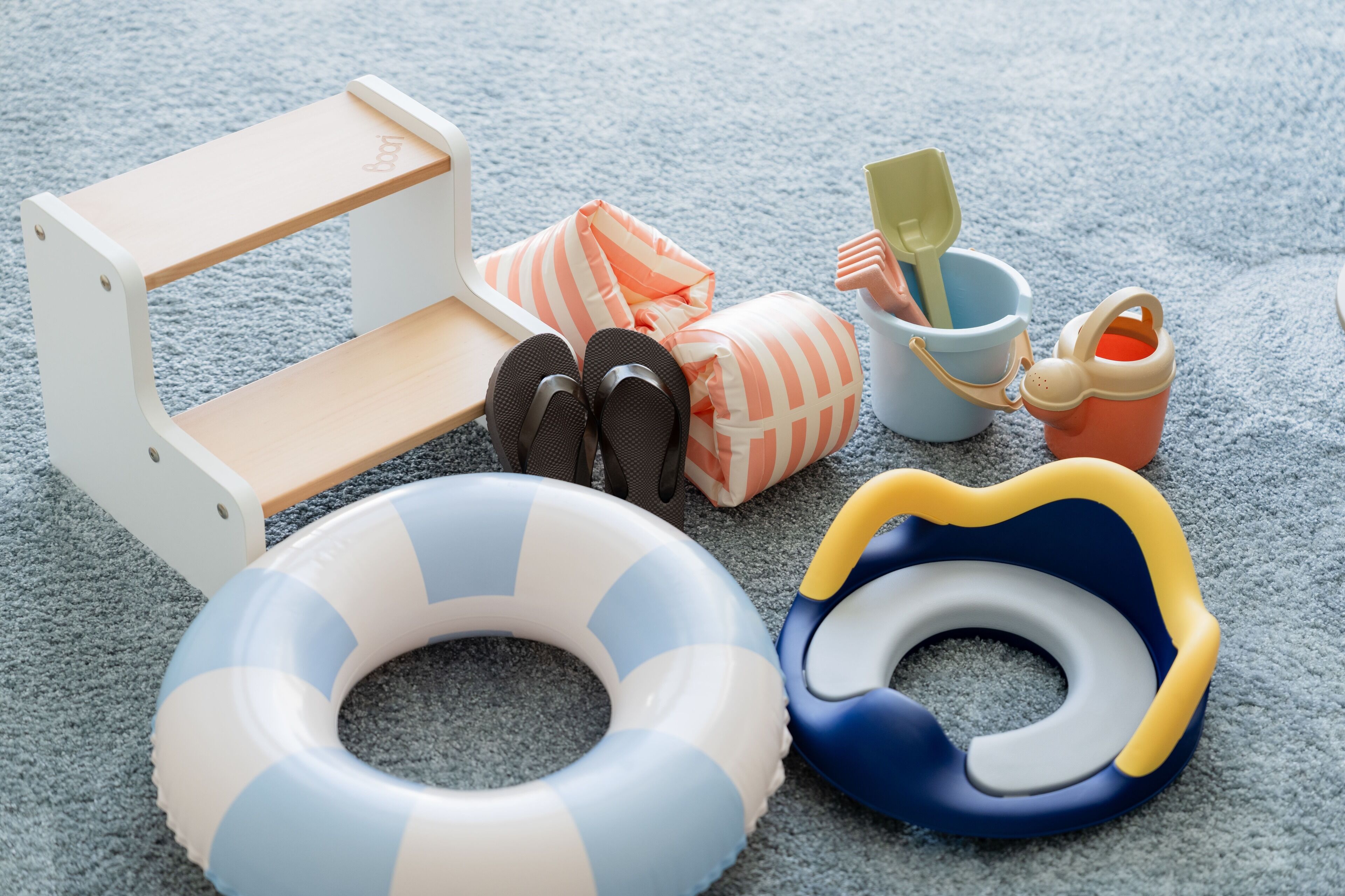 A beach-themed play area with a small wooden bench, striped pillows, a blue and white inflatable ring, a blue and yellow inflatable duck, a small bucket, and a pair of black sandals.