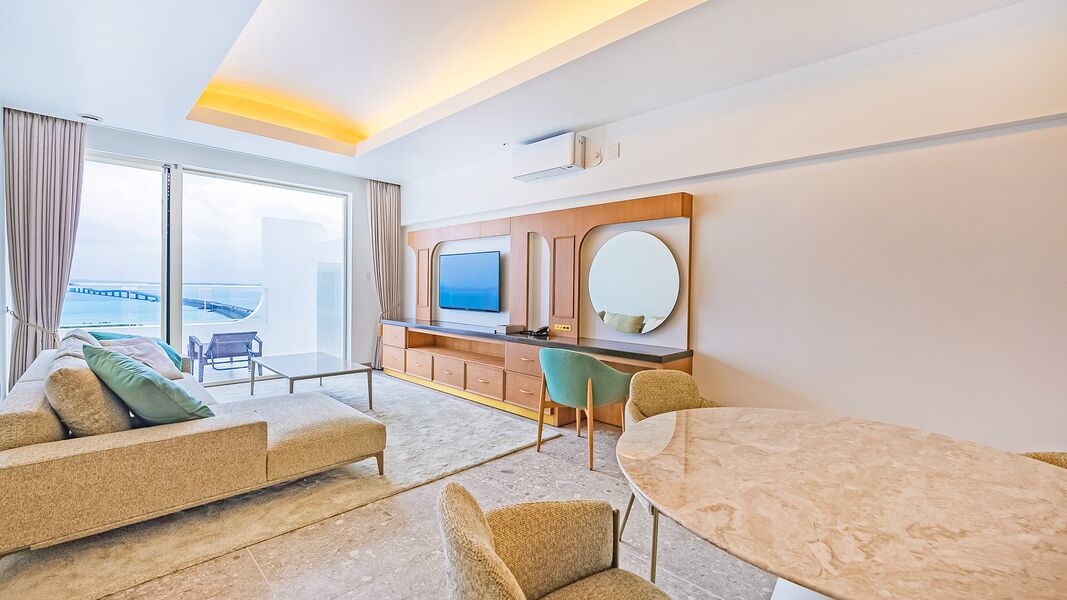 Hotelkamer met een bank, een eettafel, een televisie en een groot raam met uitzicht op een brug.