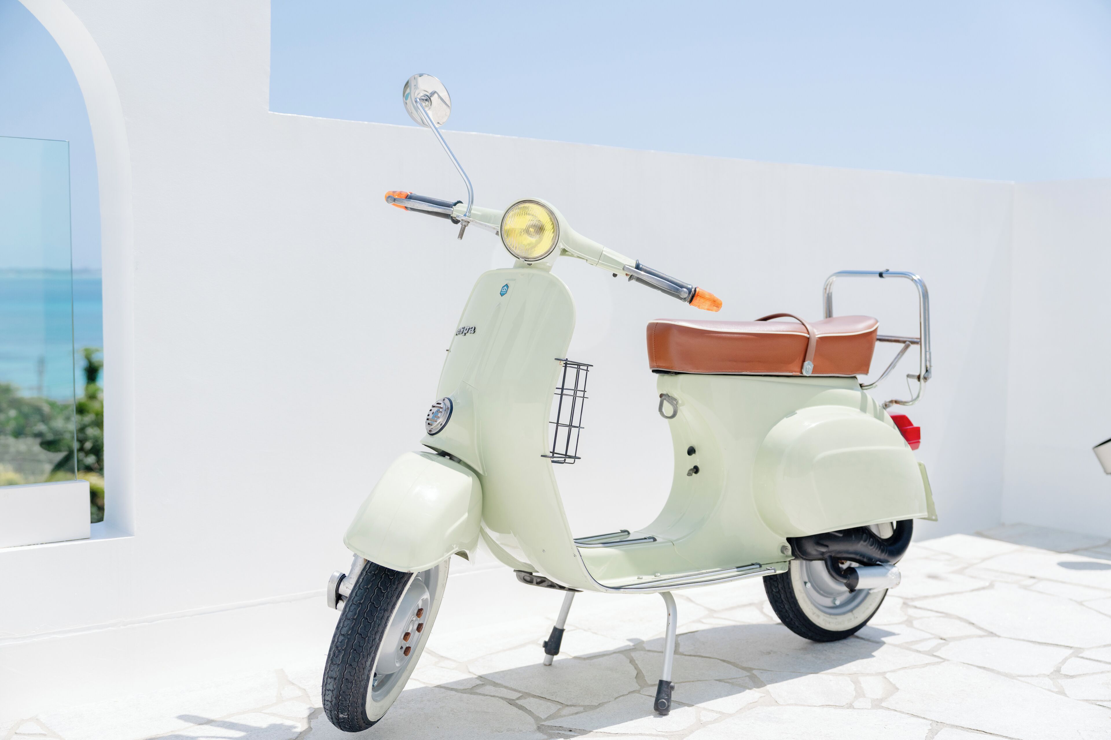 A vintage white scooter with a brown seat and chrome details, parked on a tiled patio with a white wall and a clear sky in the background.