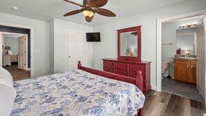 Un dormitorio con cama, cómoda roja, ventilador de techo, televisor y baño visible a través de una puerta abierta.