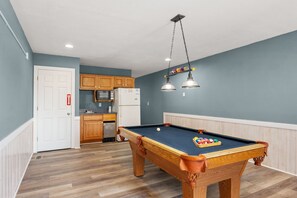 A pool table with balls, a microwave, and wooden cabinets in a room with a light blue wall and wooden flooring. - Sea You Soon: Semi-Oceanfront, Private Pool, Dog Friendly, Hot Tub and Game Room! (Southern Shores)