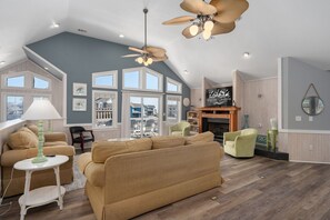A spacious living room with a large sofa, a coffee table, and a TV. There are multiple windows and a ceiling fan. - Sea You Soon: Semi-Oceanfront, Private Pool, Dog Friendly, Hot Tub and Game Room! (Southern Shores)