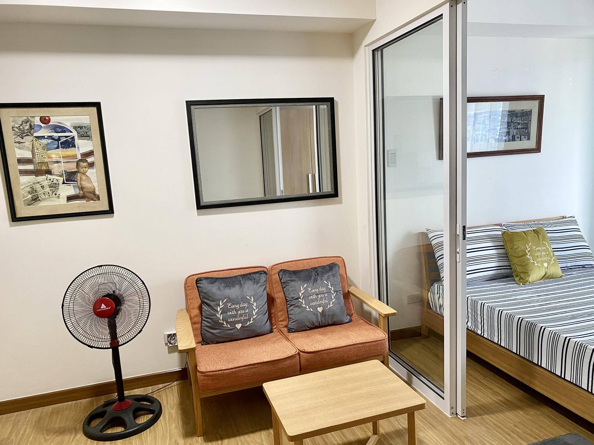A cozy living room with a sofa, a small table, a standing fan, and a bed with striped bedding.