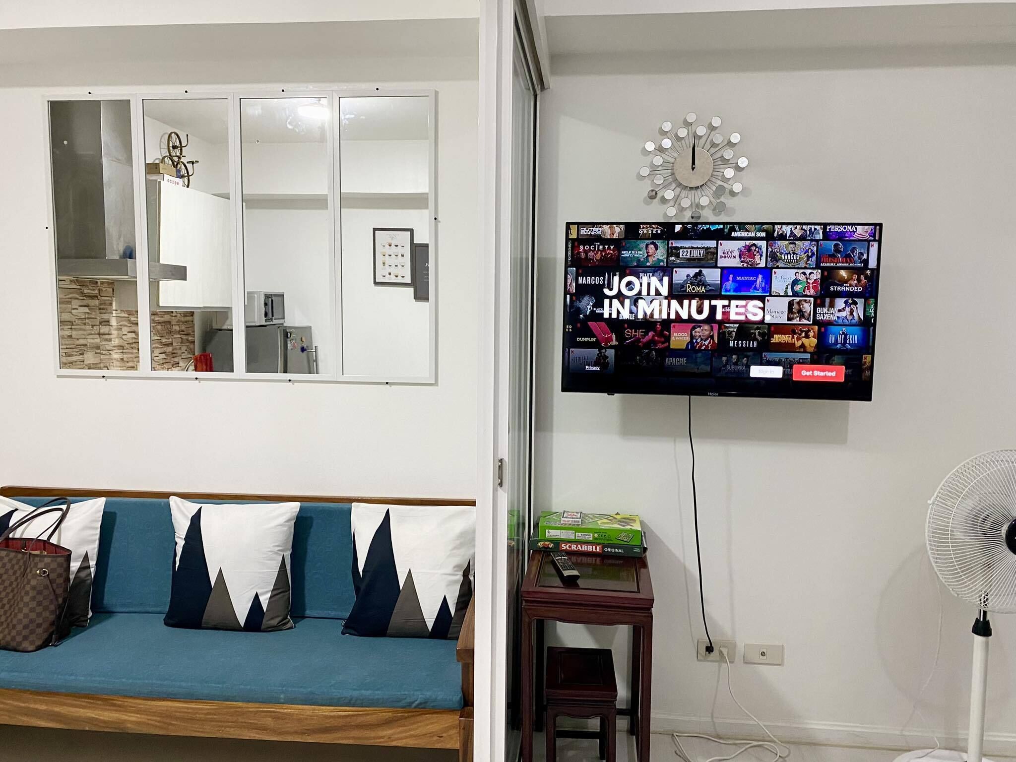 A living room with a TV displaying a gaming channel, a sofa with patterned cushions, a small table with a Scrabble box, and a wall-mounted clock.