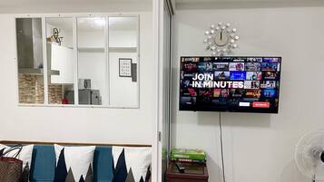 A living room with a TV displaying a gaming channel, a sofa with patterned cushions, a small table with a Scrabble box, and a wall-mounted clock.