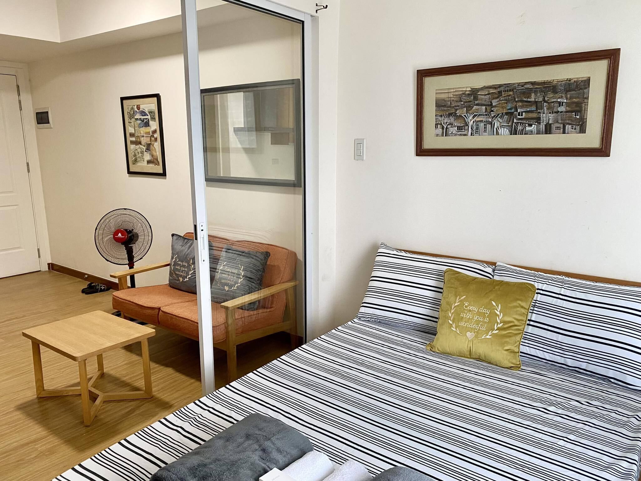 Een kamer met een bed, een stoel, een klein tafeltje en een ventilator.