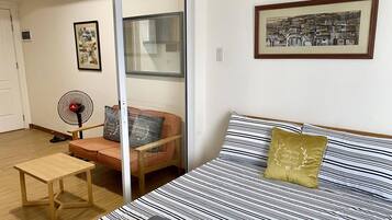 Een kamer met een bed, een stoel, een klein tafeltje en een ventilator.