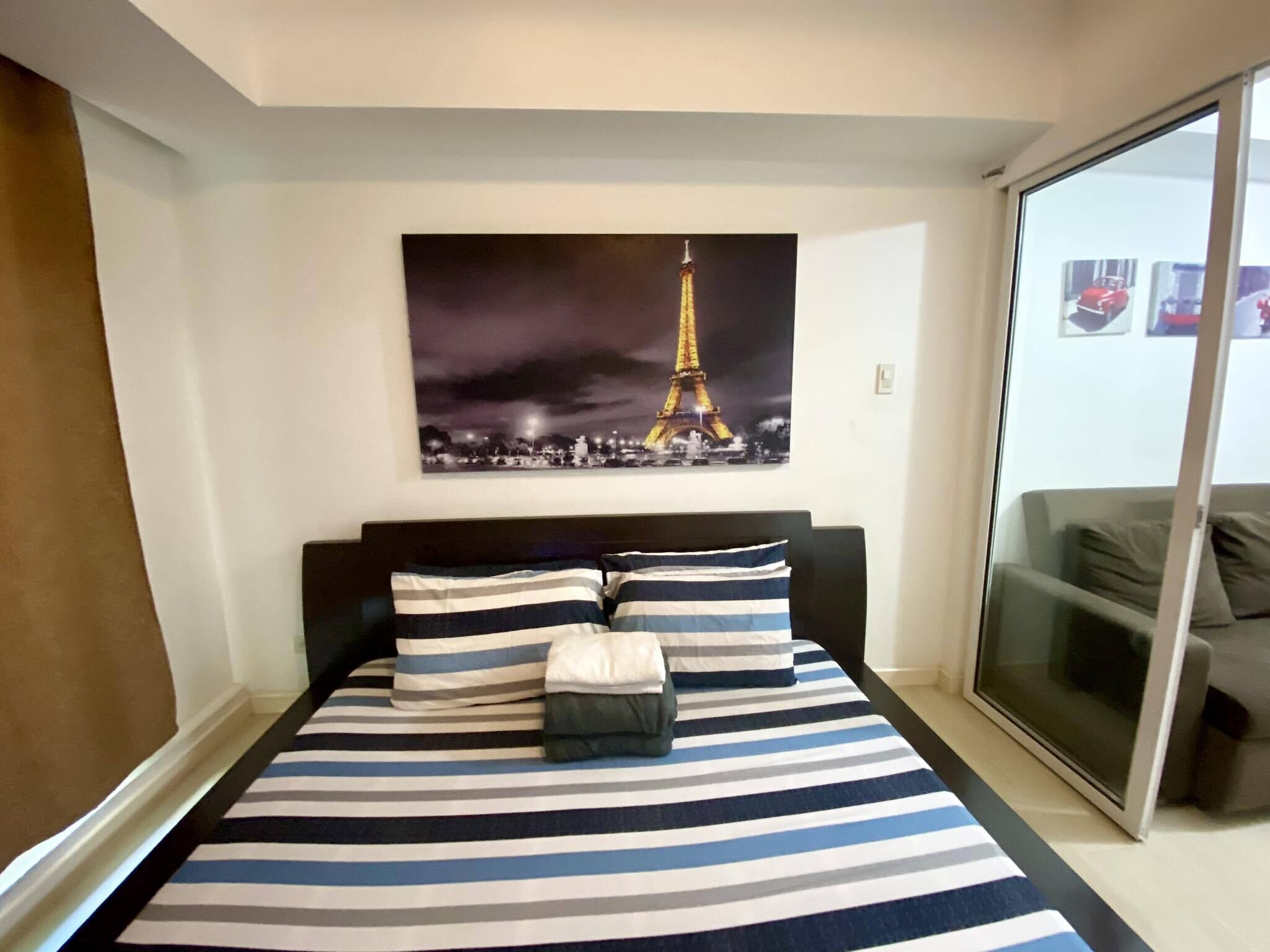 Une chambre à coucher avec un lit doté de draps à rayures, une grande fenêtre et un tableau représentant la Tour Eiffel accroché au mur.