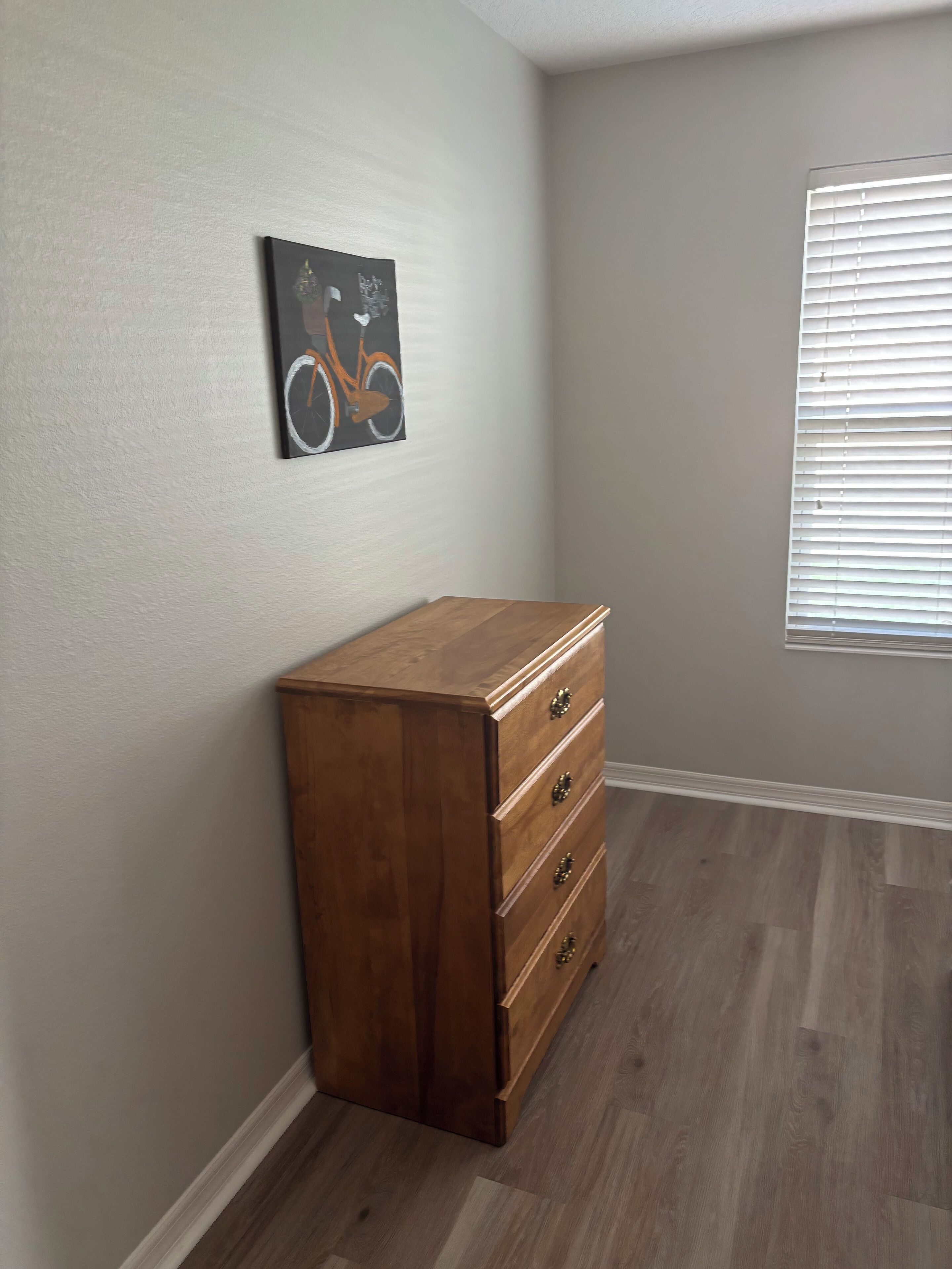 A room with a wooden dresser, a wall-mounted picture, and a window with blinds.