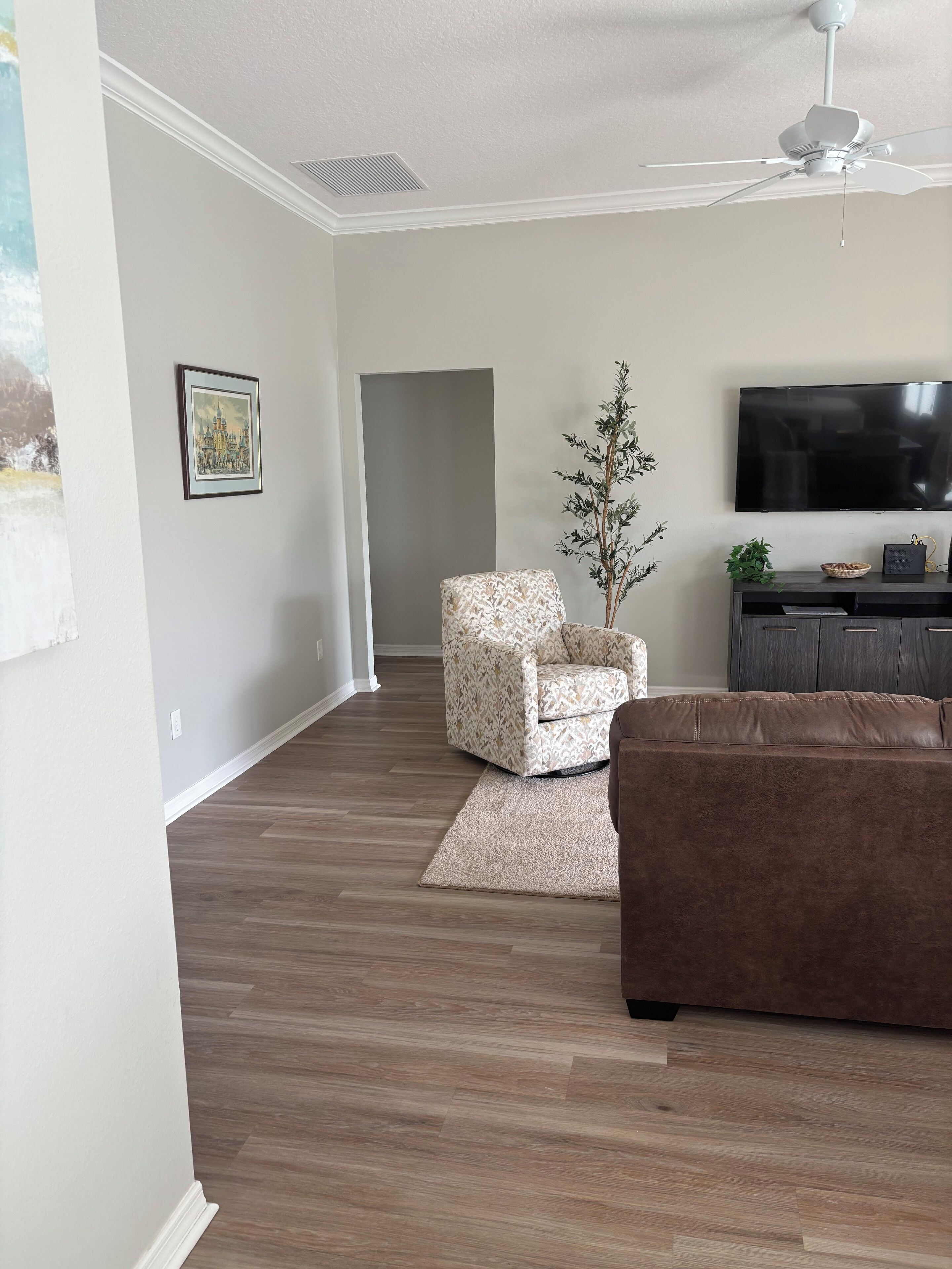 A living room with a brown couch, a patterned armchair, a small plant, a flat-screen TV, and a ceiling fan.
