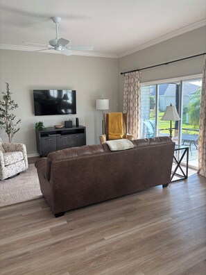 A living room with a brown sofa, a TV, a ceiling fan, a lamp, and a view of the outdoors through large windows.