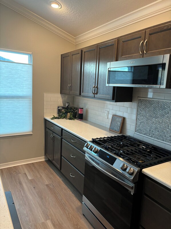 A kitchen with dark wooden cabinets, a stainless steel stove, a microwave, and a window with blinds.