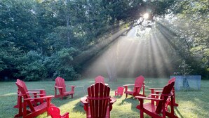 Un espace extérieur verdoyant, avec des chaises Adirondack rouges disposées autour d’une table centrale, le tout entouré d’arbres et baigné de lumière solaire.
