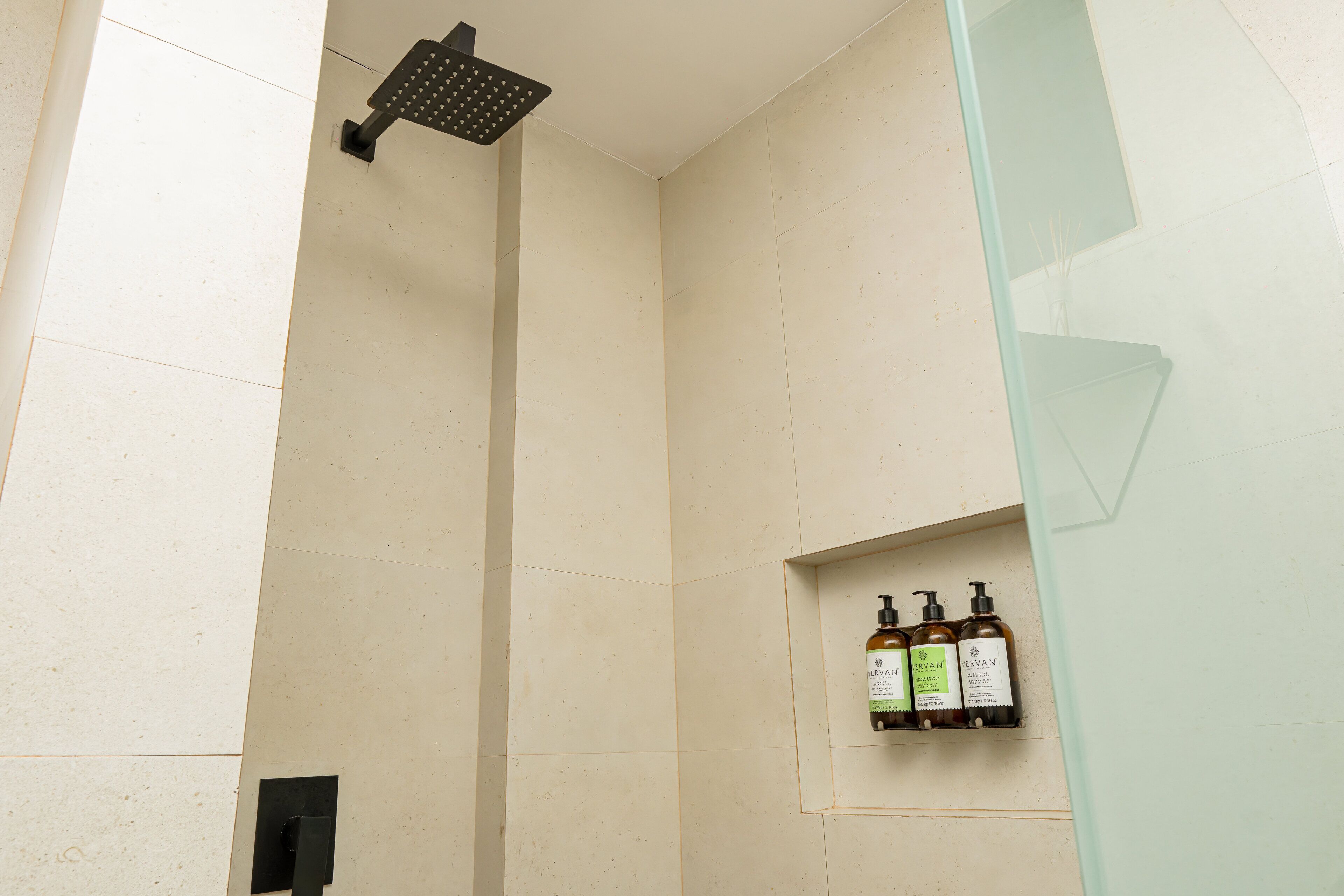 Une douche équipée d’un pommeau de douche à main et d’une étagère contenant trois bouteilles de gel douche CERVAN.
