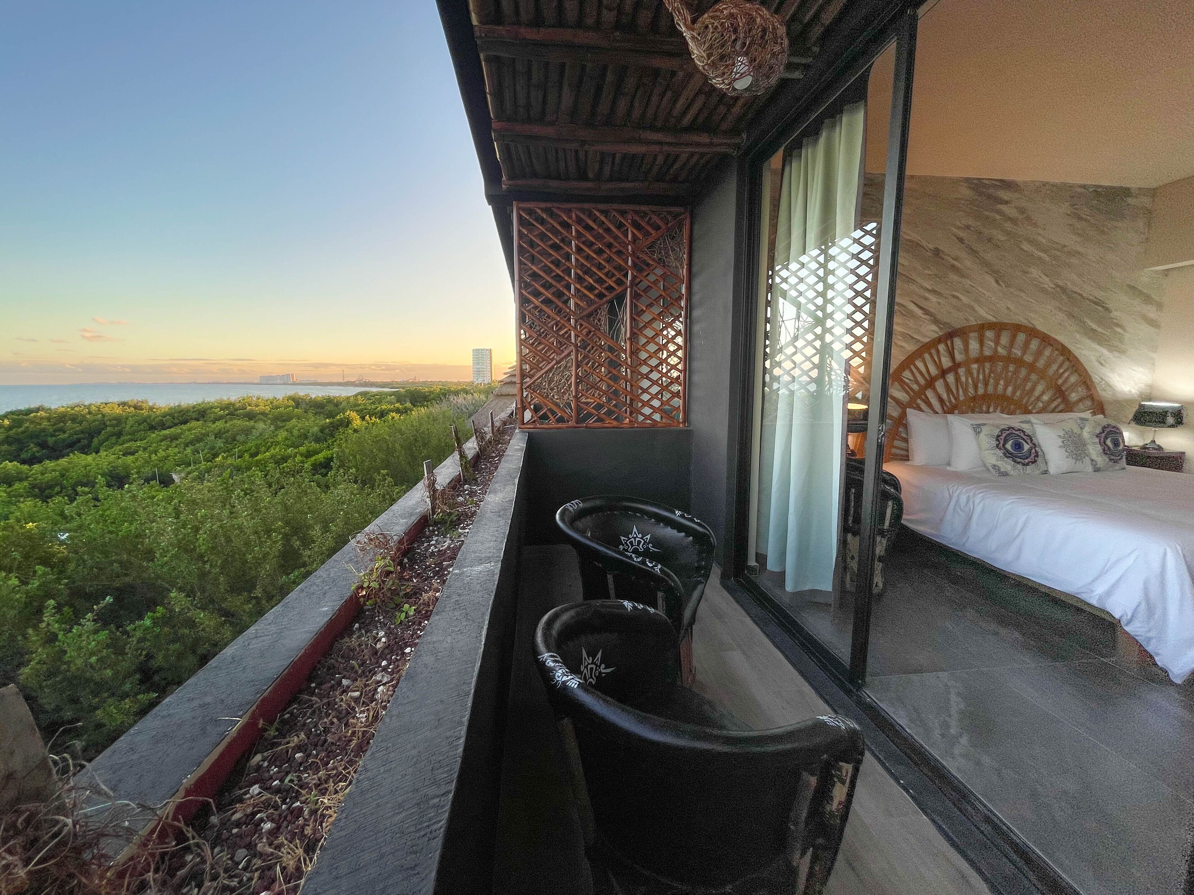 A balcony with a view of greenery and a cityscape. There is a bed with a headboard and pillows, a small table, and a chair.
