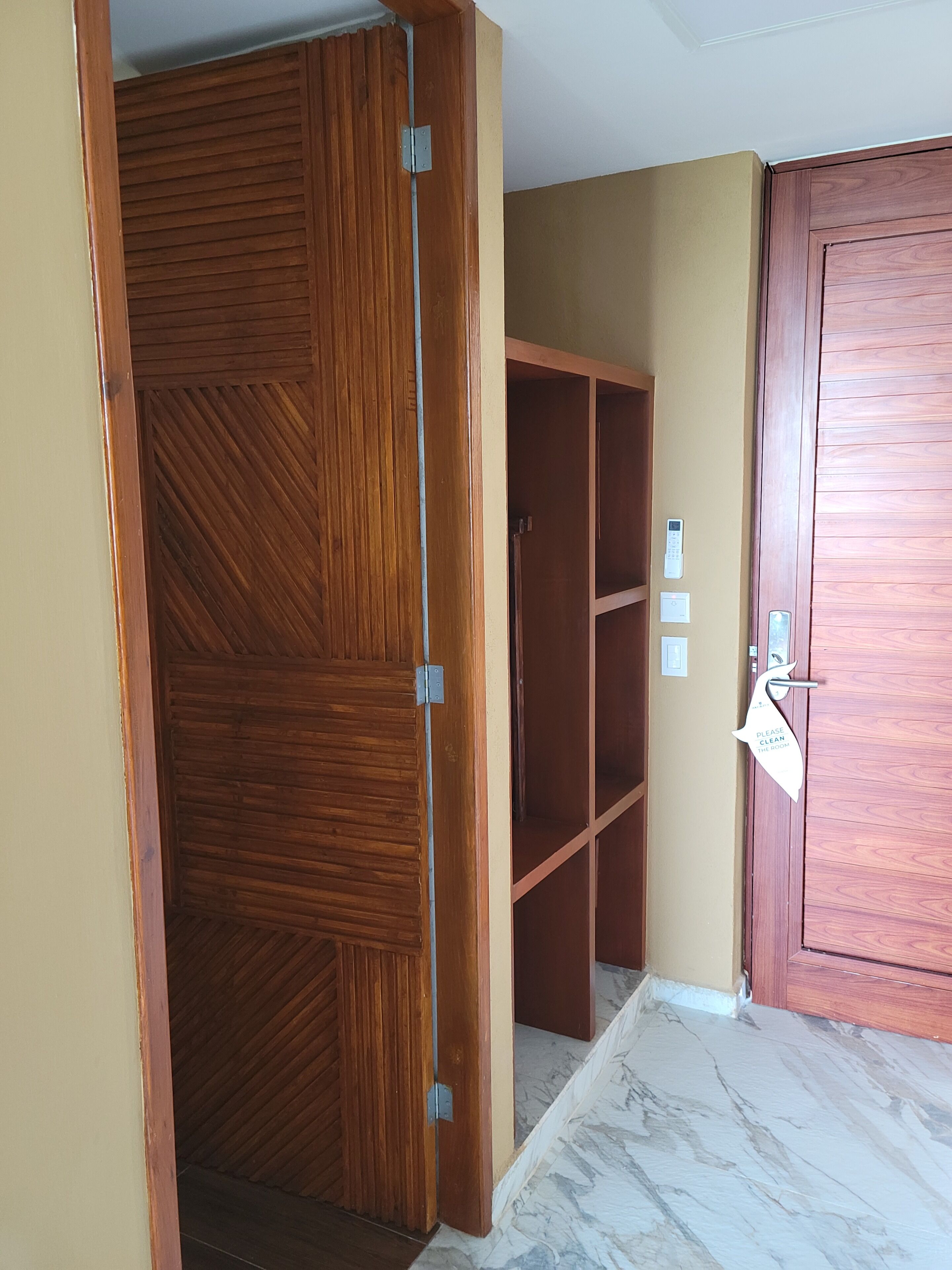 A wooden wardrobe with a mirrored door, a small shelf unit, and a door with a 'Please clean the room' sign.