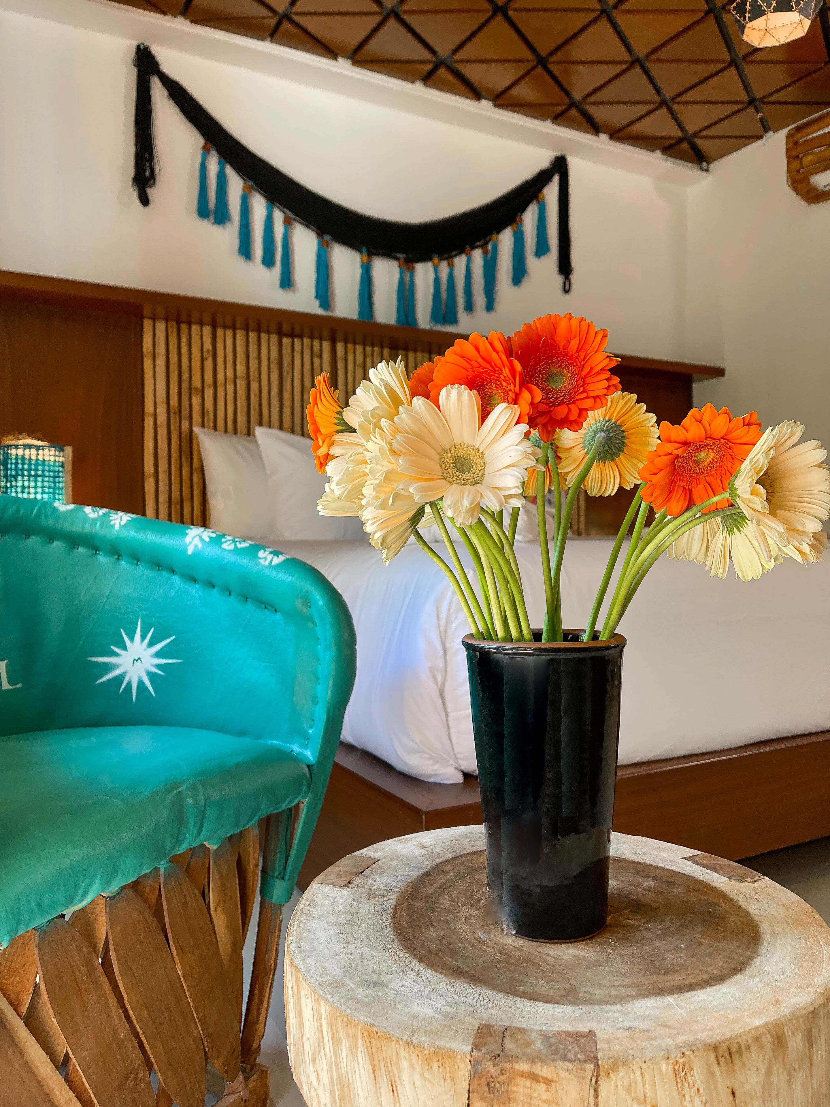 Una habitación con una cama, un cabecero de madera, una silla color aguamarina y un florero con flores sobre una mesa de madera.