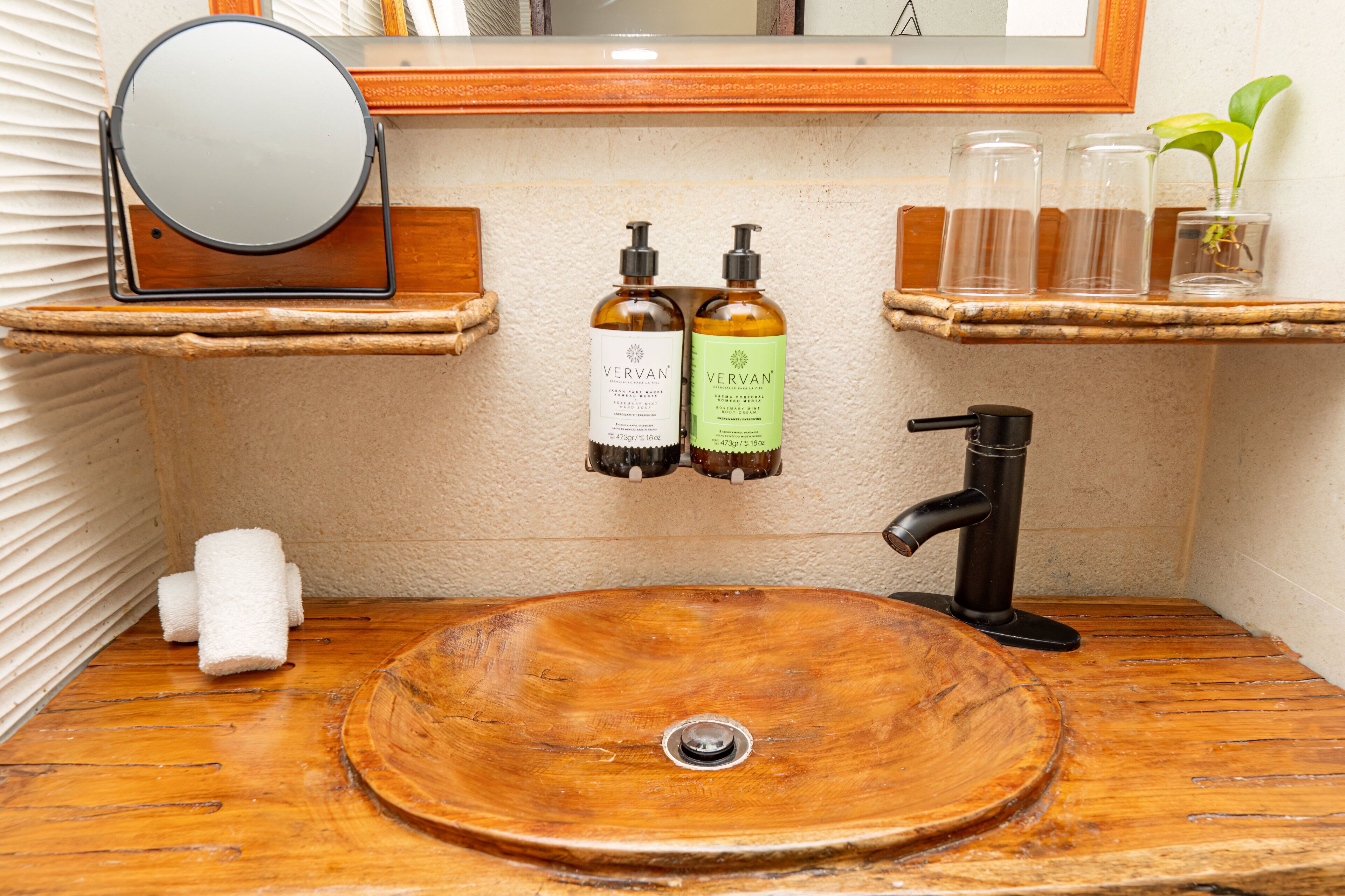 A wooden vanity with a round sink, two bottles of Vervan shampoo, a mirror, and a small plant.