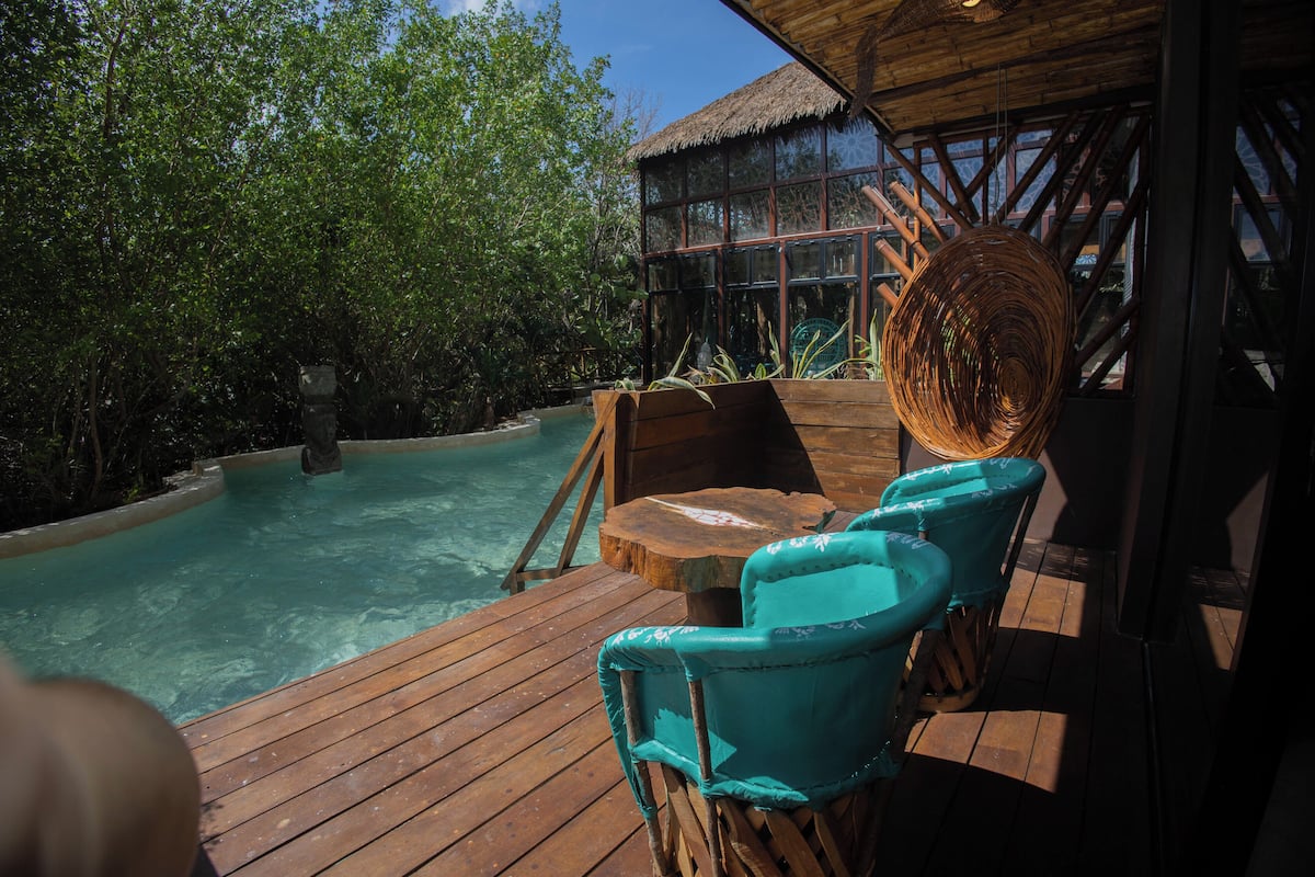 Een houten terras met een zwembad, turquoise stoelen en een rieten dakconstructie.