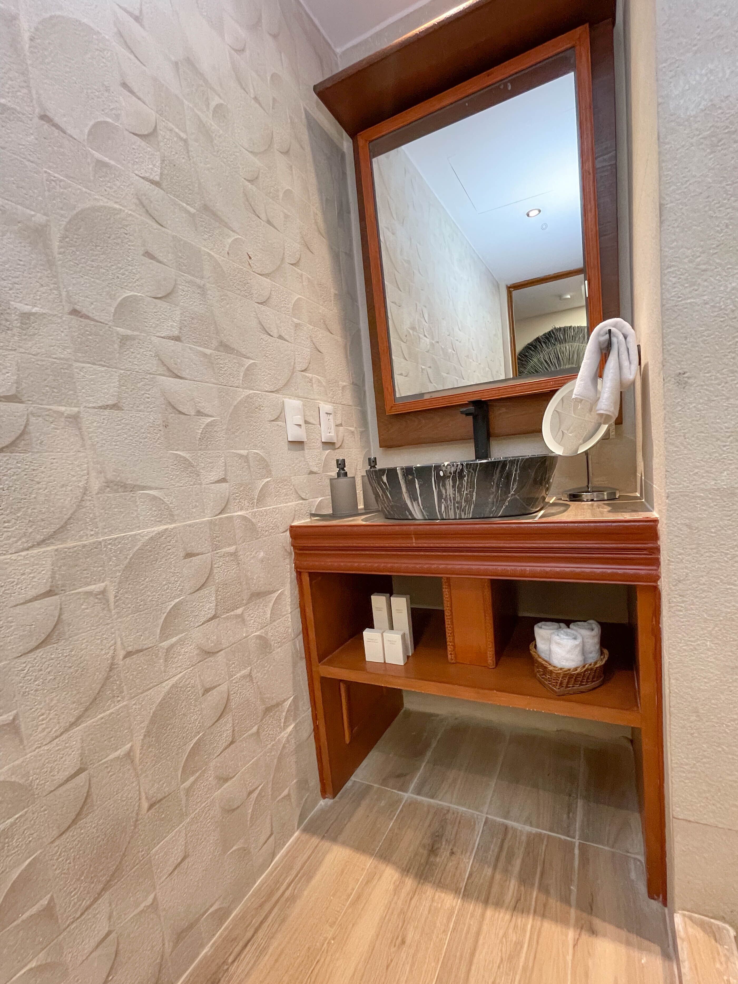 Un baño con lavamanos de mármol, tocador de madera y pared con textura.