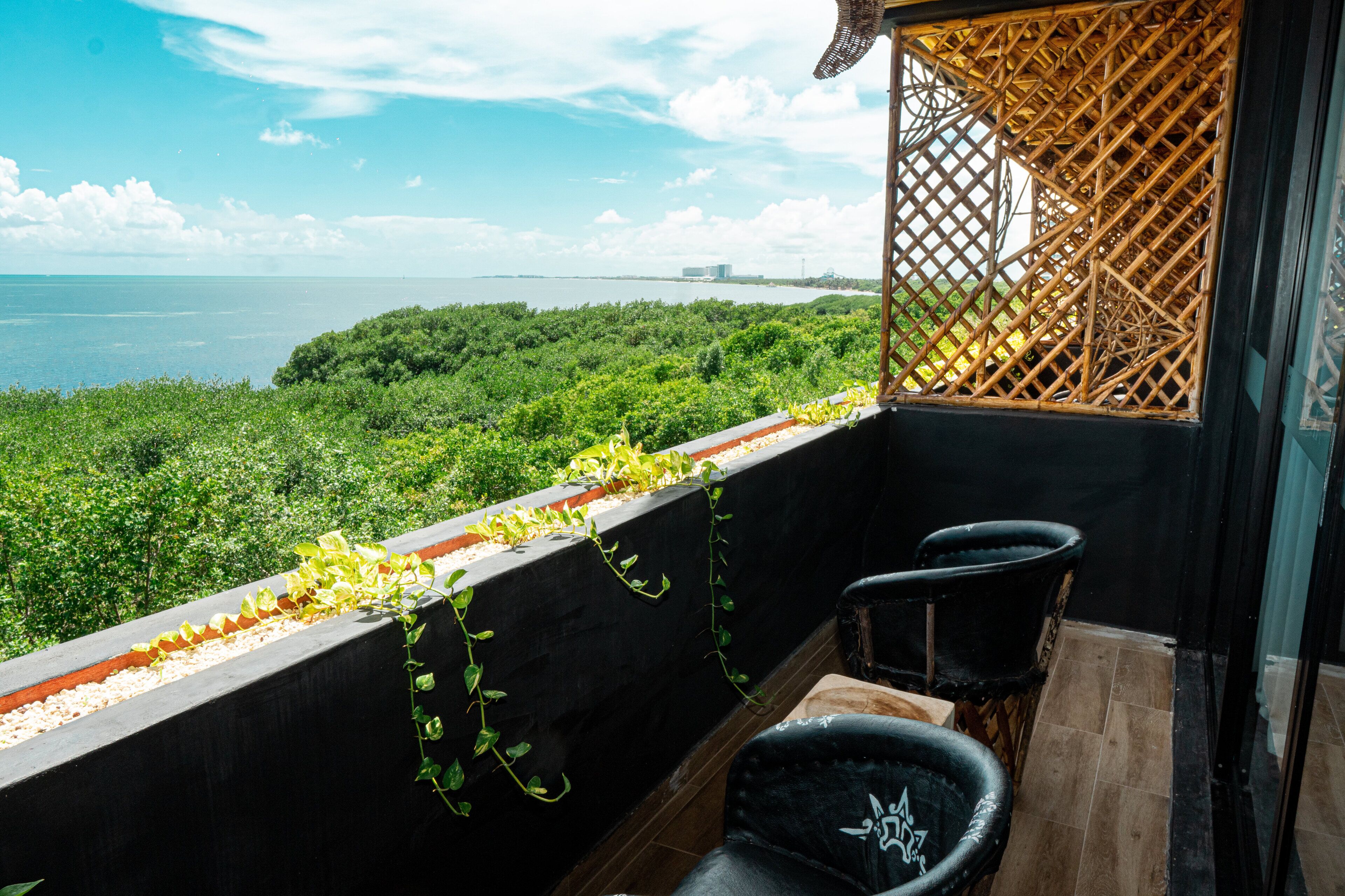A balcony with a view of the sea and greenery, featuring two black chairs and a woven partition.