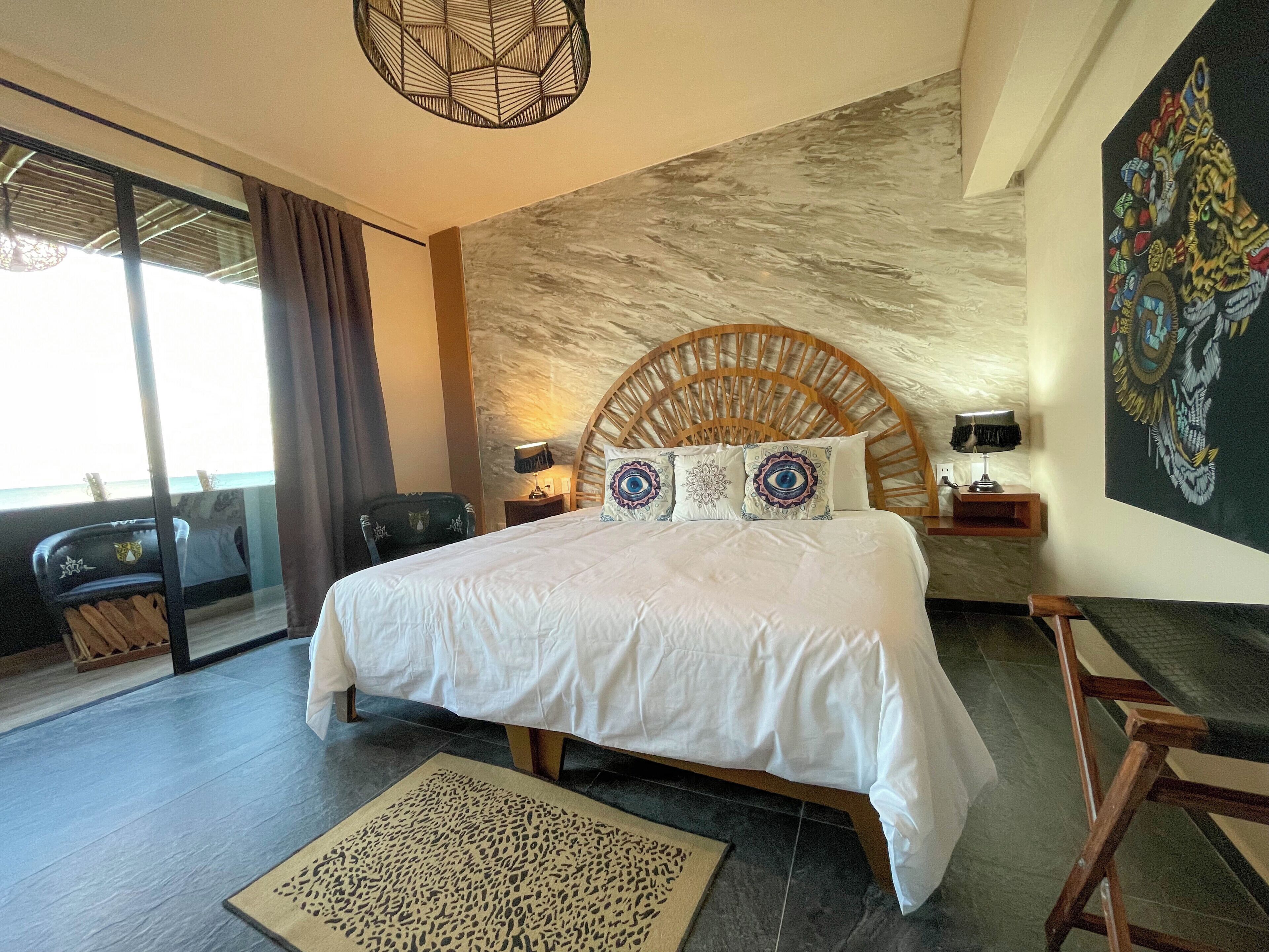 Habitación de hotel con cama, cabecera, mesita de noche, lámpara, sillón, ventana grande y un cuadro en la pared.