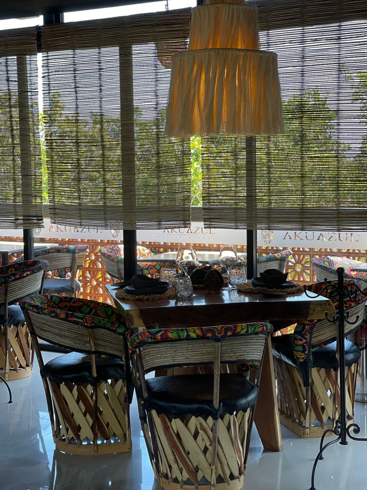 A dining area with a table set for four, surrounded by patterned chairs, a hanging light fixture, and a view through large windows with blinds.