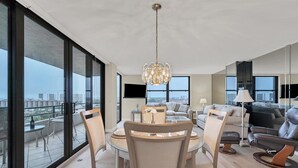 A spacious living area with a dining table, chairs, and a chandelier. Large windows offer a view of the cityscape.