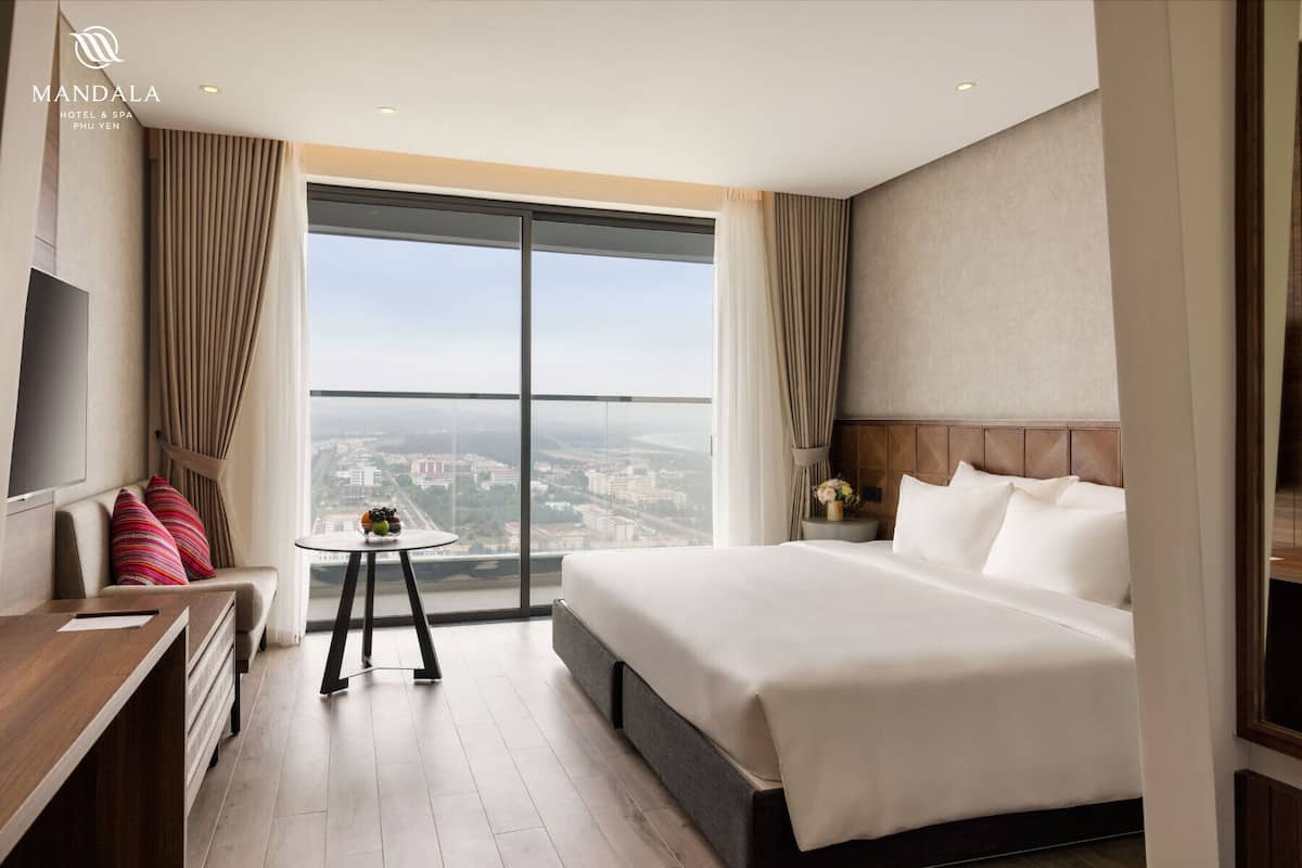 A hotel room with a large bed, a desk, a small table with fruit, and a view of the city through large windows.
