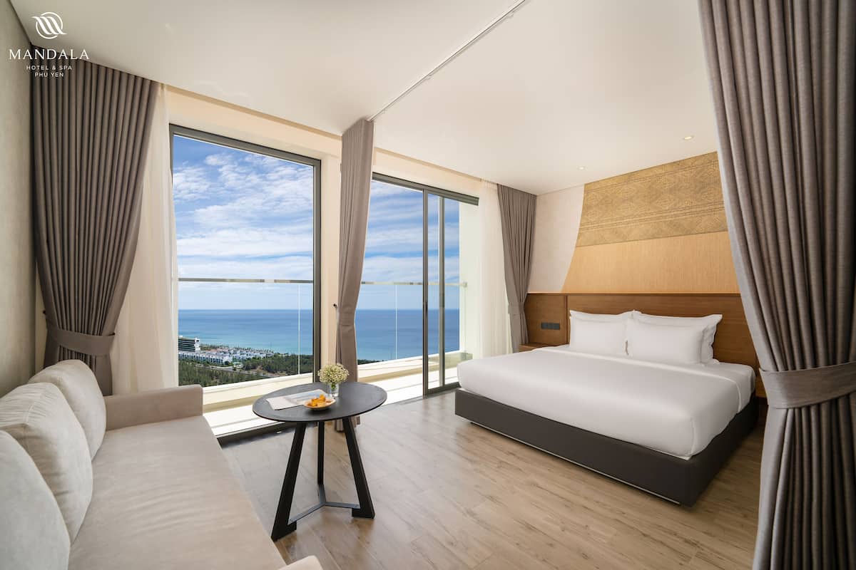 A modern hotel room with a large bed, a sofa, a small table with a vase, and a view of the sea.