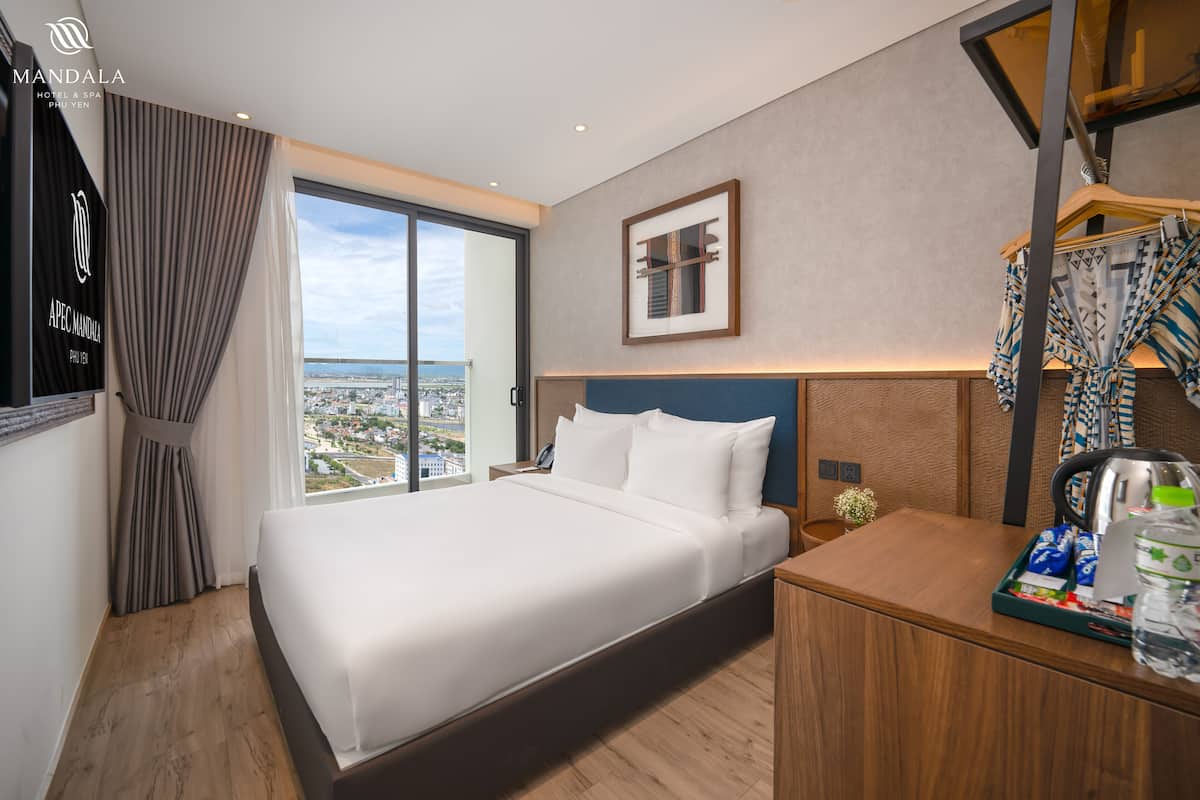 A hotel room with a large bed, a desk with a tray of toiletries, a framed picture on the wall, and a view of the city through a large window.