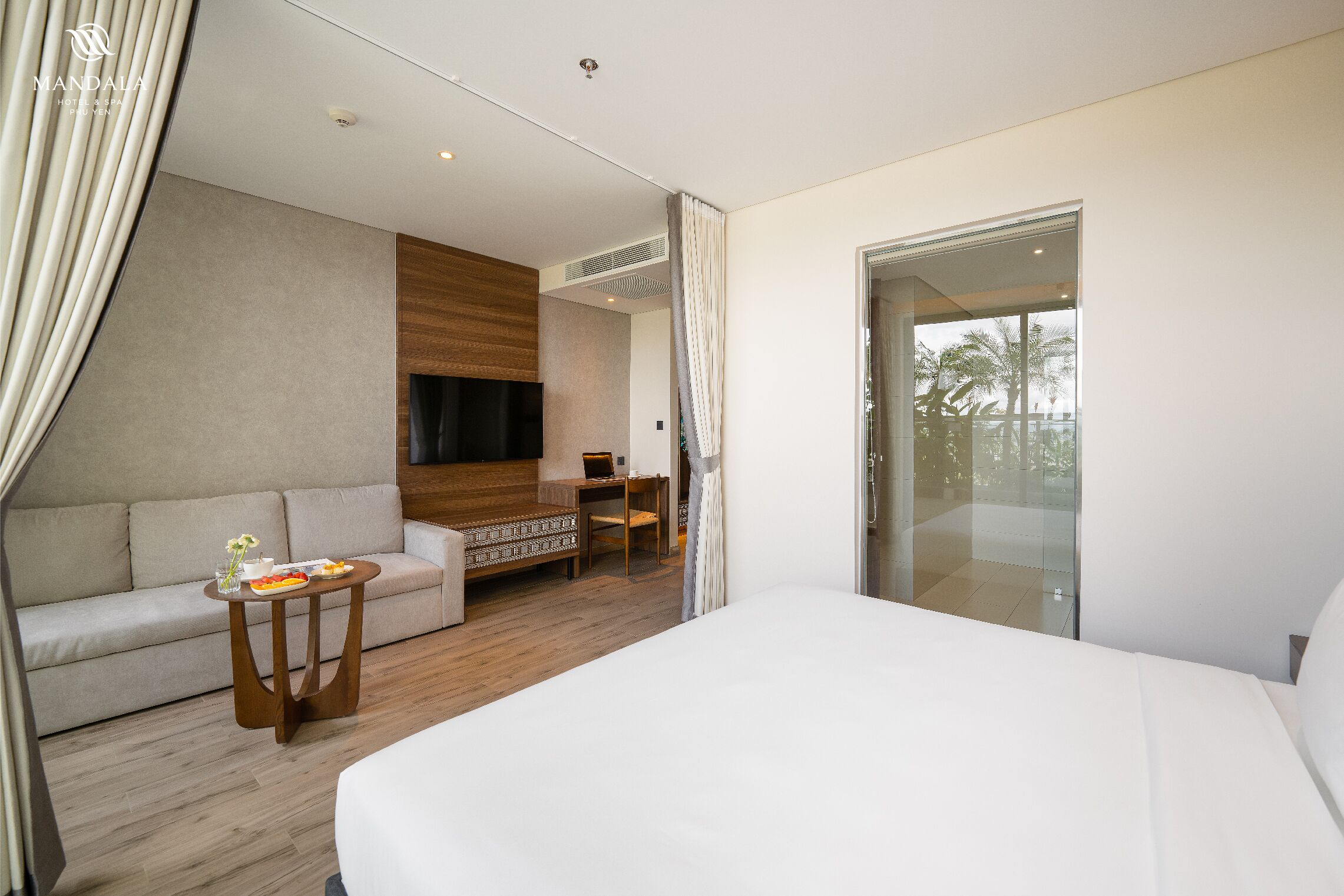 A modern hotel room with a large bed, a sofa, a small table with a plate of food, and a TV on the wall.