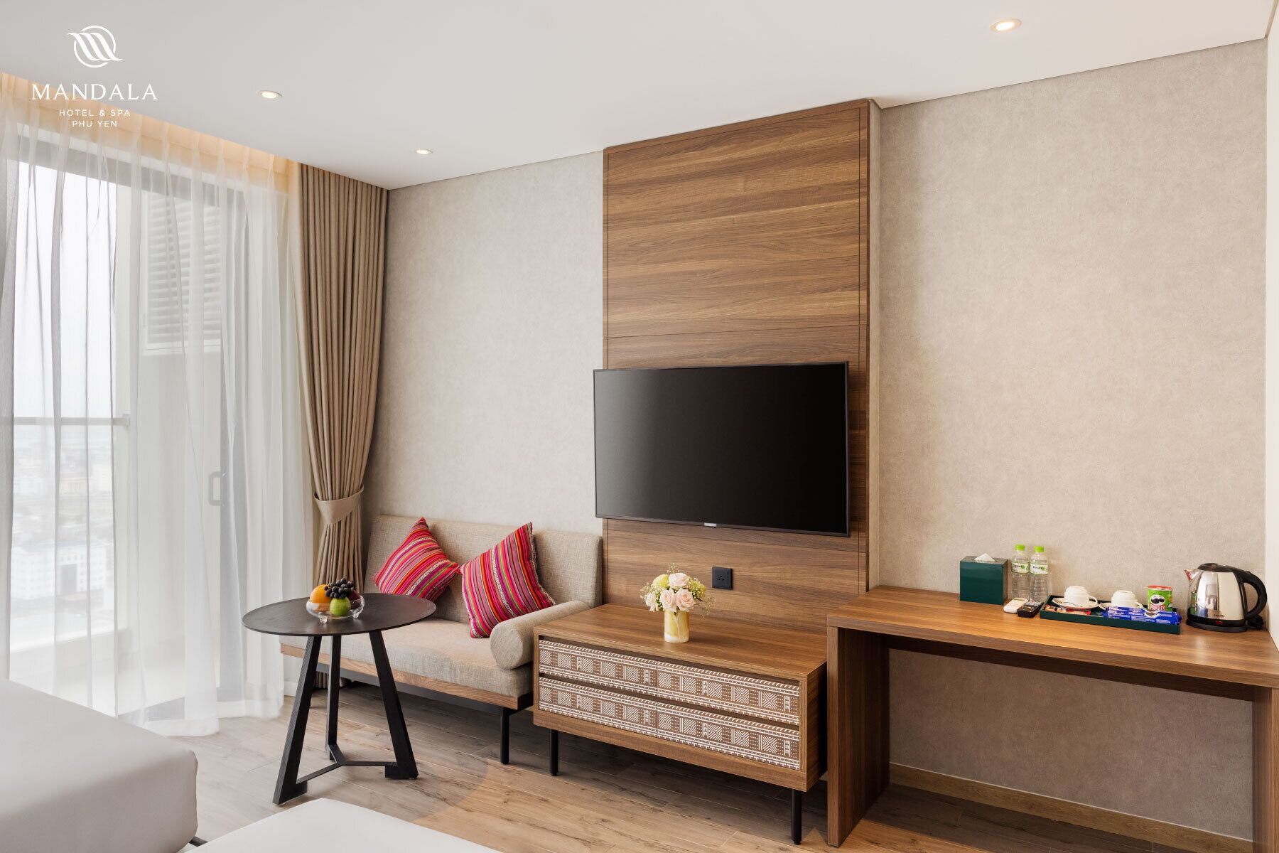 A modern hotel room with a large flat-screen TV mounted on a wooden panel wall, a sofa with colorful cushions, a small round table with fruit, and a coffee station.