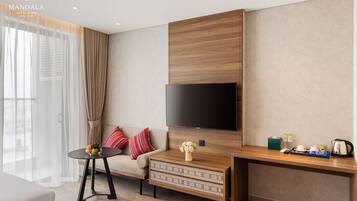 A modern hotel room with a large flat-screen TV mounted on a wooden panel wall, a sofa with colorful cushions, a small round table with fruit, and a coffee station.