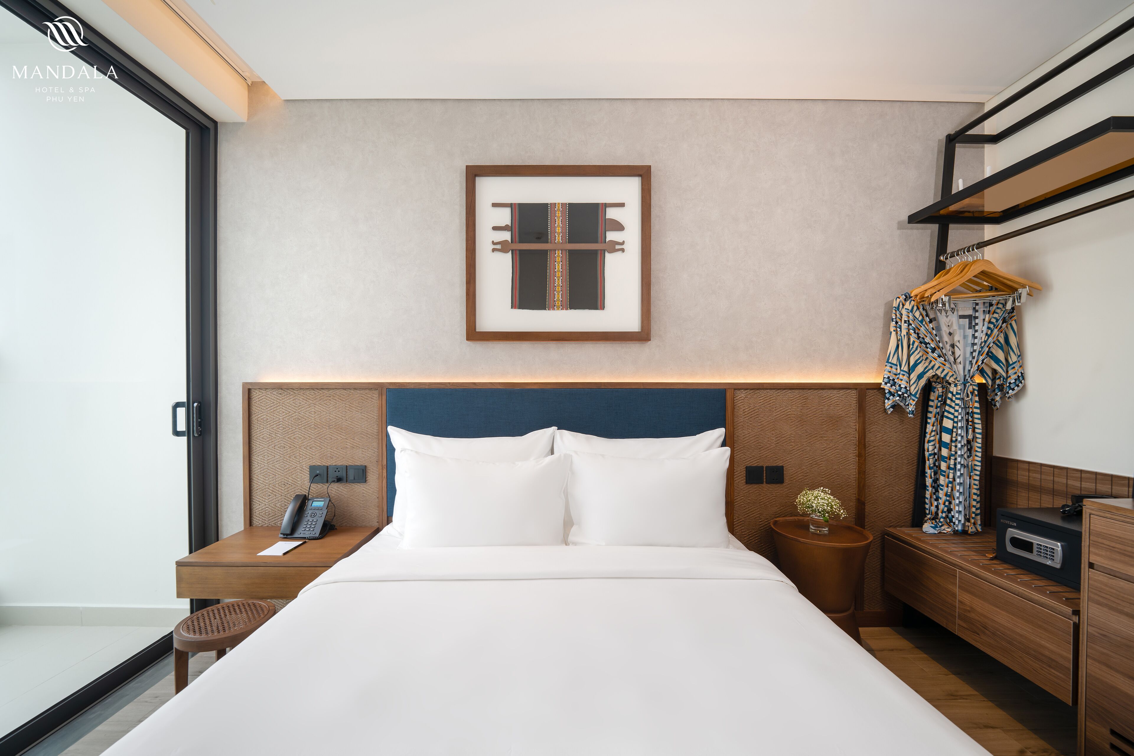 Ein Hotelzimmer mit einem großen Bett, einem Nachttisch mit Telefon, einem kleinen Tisch mit einer Vase und einem Kleiderständer mit einem gemusterten Kleidungsstück.